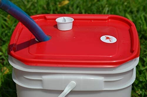 Red-lid bucket with a hose inserted on grassy ground.